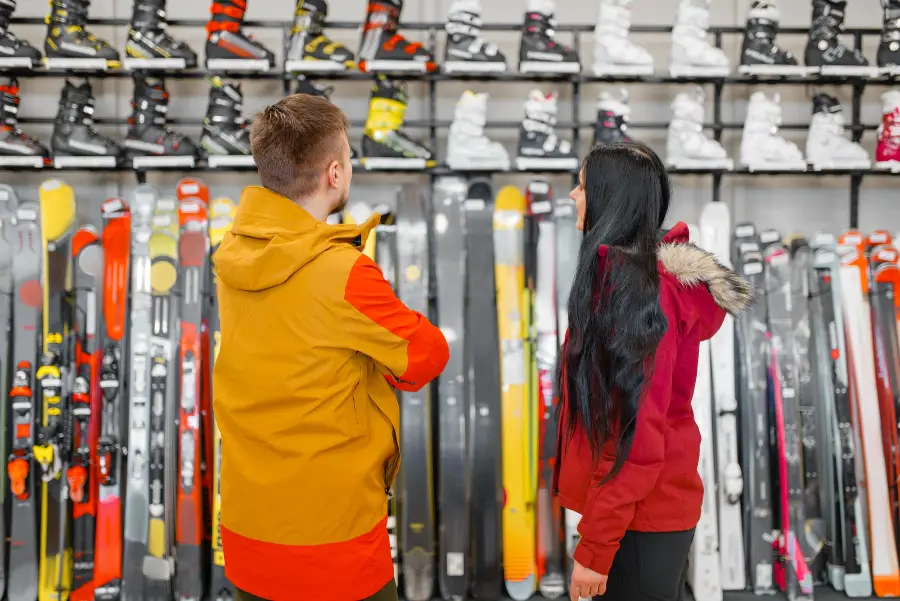 Two customers, a man in a yellow jacket and a woman in a red coat, browsing a variety of skis and boots displayed on racks at Bansko Skis Rental, showcasing premium equipment and personalized service.