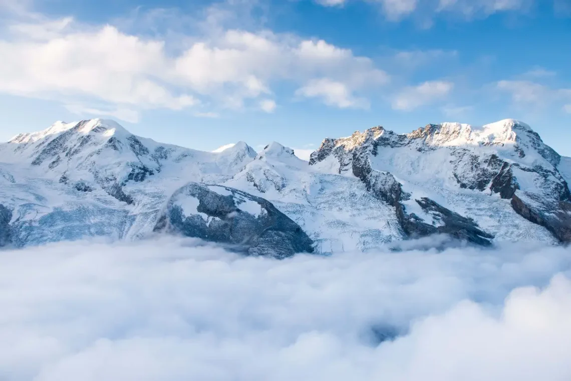 A stunning panoramic view of snow-covered mountain peaks towering above a sea of clouds, under a bright blue sky in Bansko, capturing the serene beauty of the region's alpine landscape.