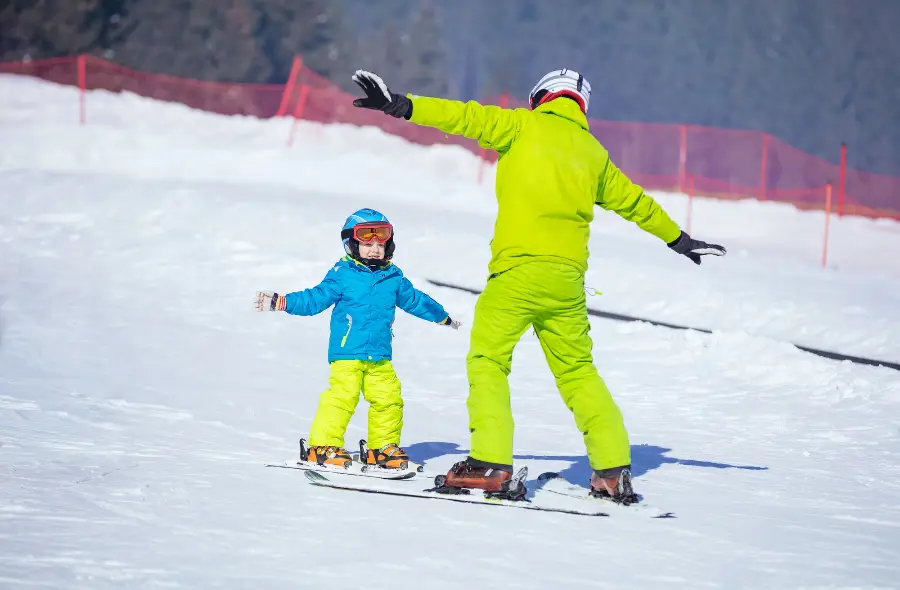 A young child in blue and neon green winter gear learning to ski with an instructor in matching green attire on a sunny ski slope in Bansko, emphasizing Bansko Skis Rental's expert ski lessons for beginners.