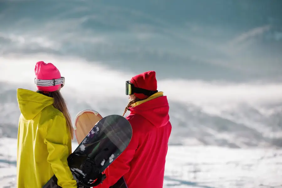 Two snowboarders in bright yellow and red winter jackets and pink hats admire the snowy mountain scenery in Bansko, carrying snowboards, showcasing Bansko Skis Rental's premium snowboard rentals and breathtaking slopes.