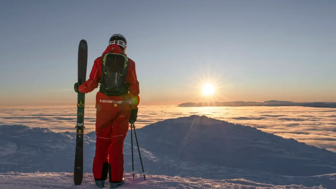 A skier in red gear with a backpack and skis stands on a snowy mountain peak, gazing at a breathtaking sunrise above the clouds in Bansko, highlighting the unforgettable alpine experiences offered by Bansko Skis Rental.